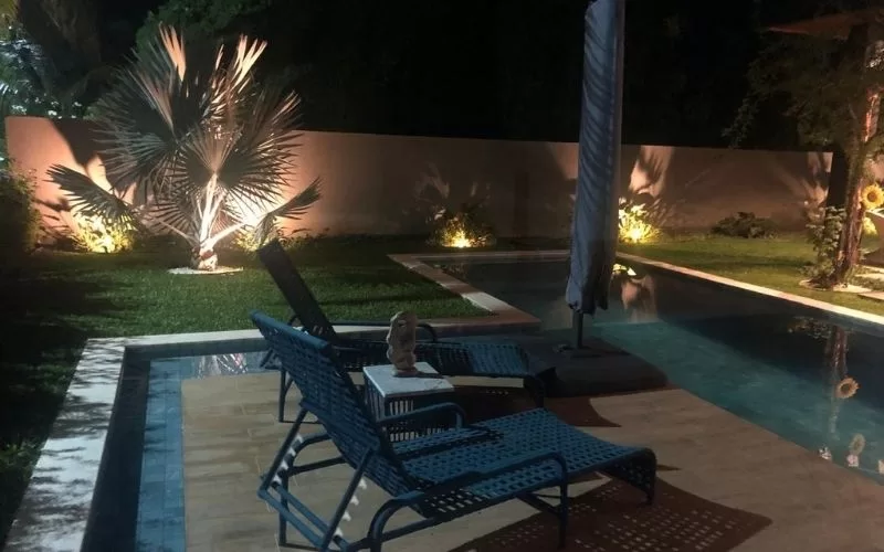 Piscina iluminada à noite com cadeiras e árvores.