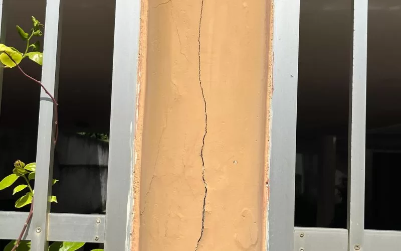 Coluna rachada entre grades cinzentas e plantas.