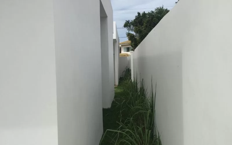Passagem estreita entre muros brancos com vegetação.